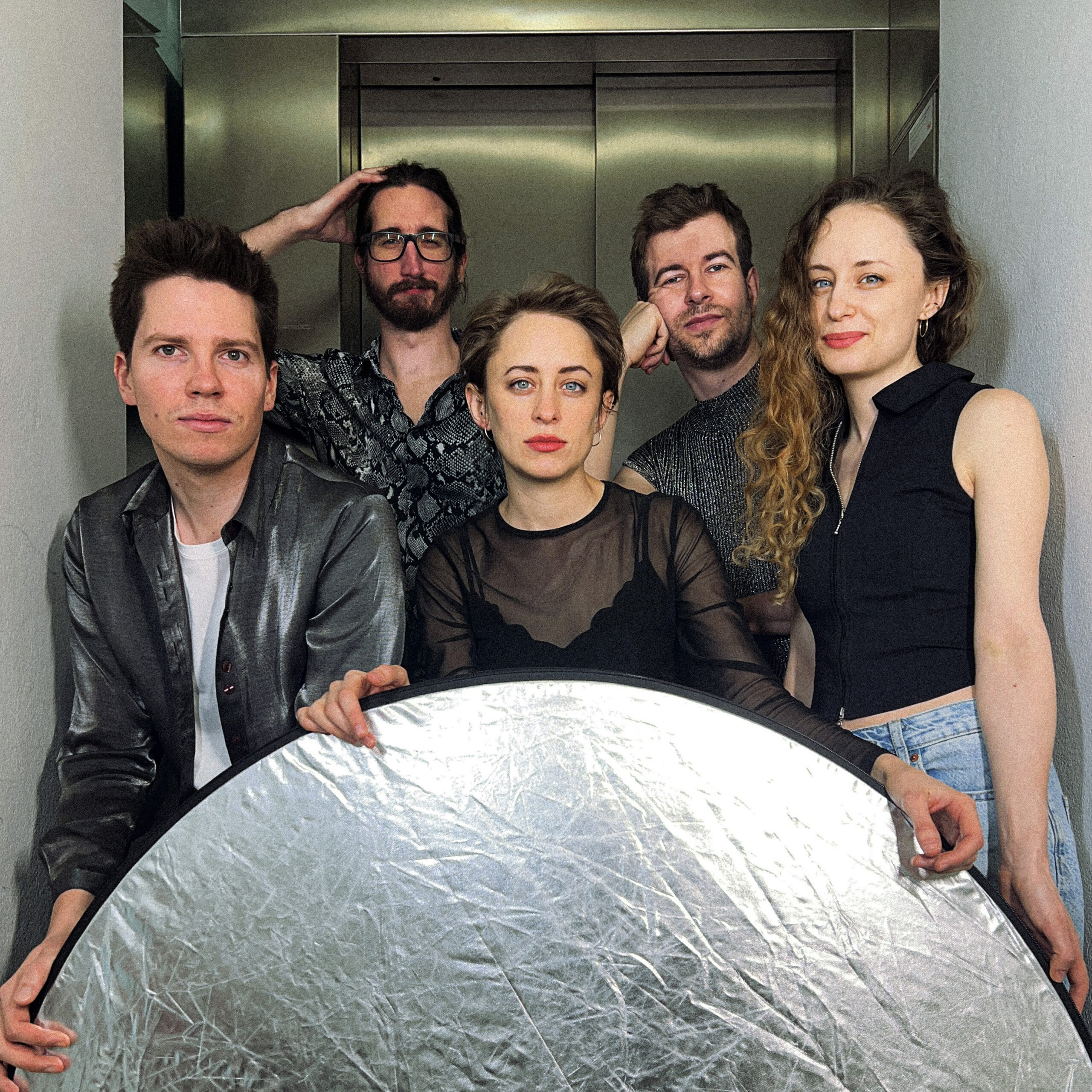 Blechkasten_Band-Quadrat Die Band Blechkasten posiert vor einem Lift. Sie besteht aus fünf Personen, zwei Frauen und drei Männer. Alle sehen direkt in die Kamera.